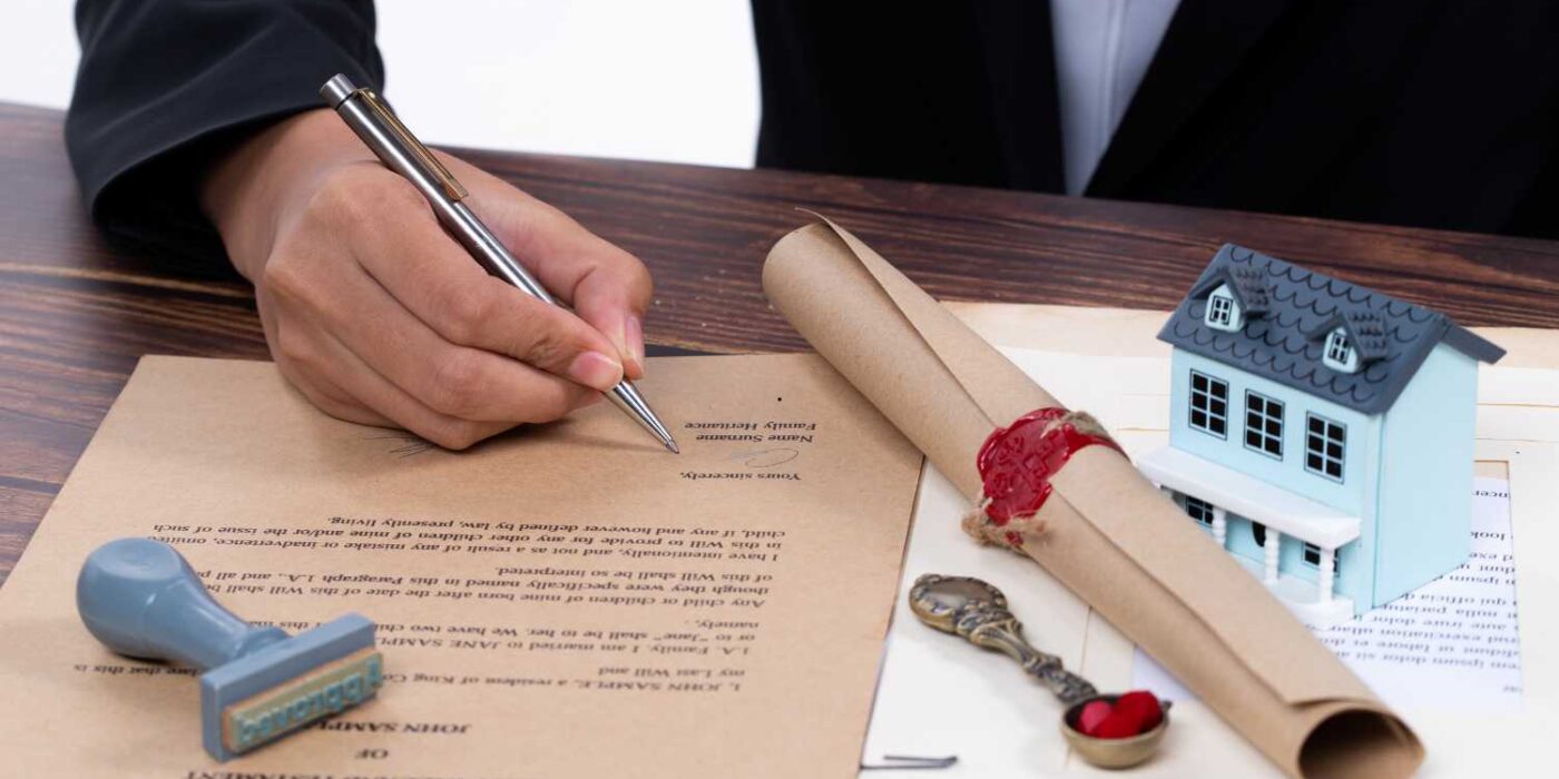 Person signing a legal document related to property inheritance, with a stamp, a rolled-up parchment sealed with red wax, and a small model house placed on a wooden desk.